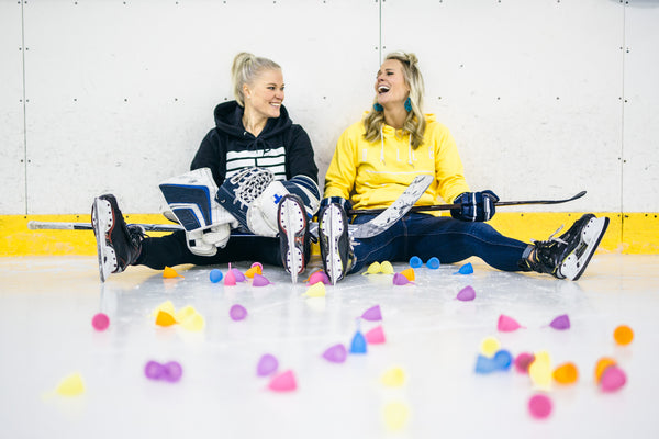 Two women ice hockey players sitting on the ice rink surrounded by menstrual cups and their ice hockey gear.